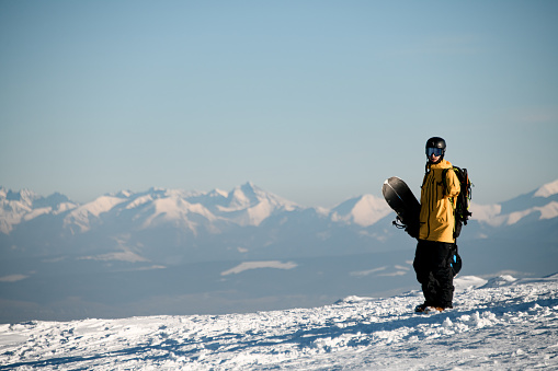 Male snowboarder standing on sunny day against backdrop of beautiful mountain peaks shrouded in fog. Young man standing outdoors in full winter sport equipment and looking at camera.