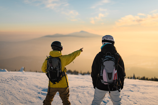 Two snowboarders enjoying beautiful natural landscape, standing on top of the mountain on sunny winter day. Man pointing to the background showing beautiful landscape