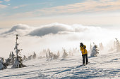 Man skier stand on the mountain top with backpack against fluffy clouds and snow covered trees