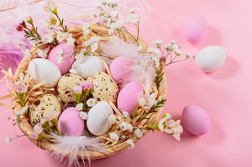 Easter candy chocolate eggs and almond sweets lying in a bird's nest decorated with flowers and feathers on pink background. Happy Easter concept.