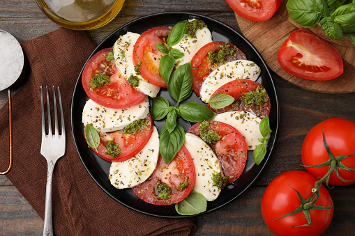 Plate of delicious Caprese salad with pesto sauce and ingredients on wooden table, flat lay