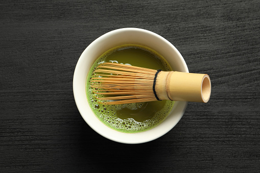 Cup of fresh matcha tea with bamboo whisk on black wooden table, top view