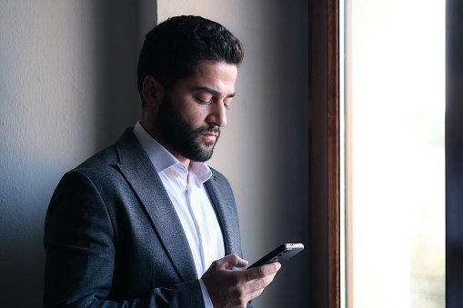 A serious man standing near the window of the office with a smartphone.
