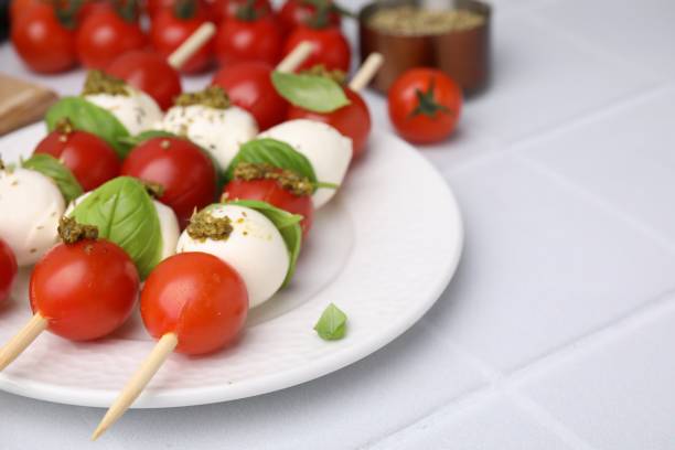 istock Caprese skewers with tomatoes, mozzarella balls, basil and pesto sauce on white tiled table, closeup. Space for text 1905398049
