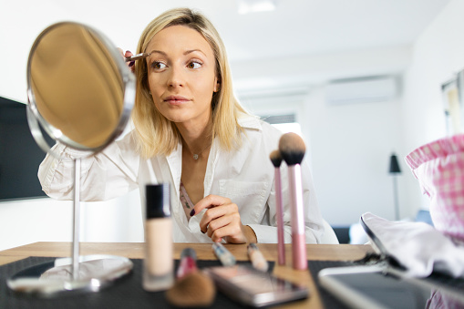 A young woman applying make up in her living room in the morning, she is sitting at a desk, and looking at herself in the mirror while using eyebrow pencil. She has a smile on her face.