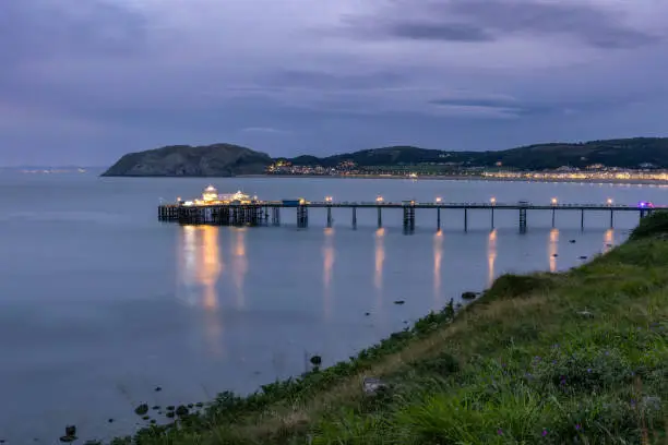 Photo of Long historic pleasure pier at dusk with lights reflecting in the water. Llandudno, UK