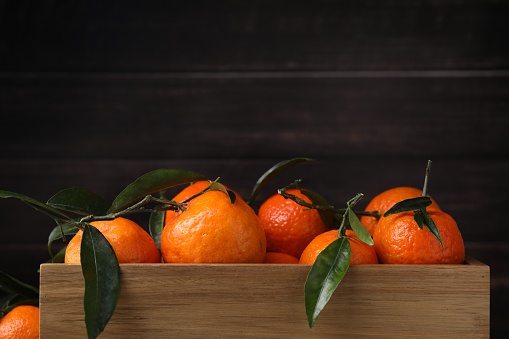 Fresh ripe tangerines with green leaves in wooden crate on dark background, closeup. Space for text