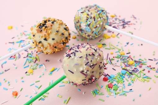 Delicious confectionery. Sweet cake pops decorated with sprinkles on pale pink background, closeup
