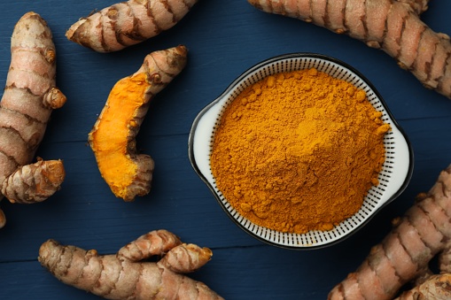 Bowl with turmeric powder and raw roots on blue wooden table, flat lay