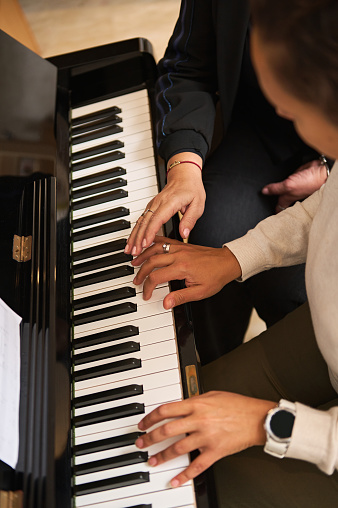 Overhead view of a young woman learning playing piano, touching piano keys under the guidance of her teacher during individual music lesson indoor. Art, culture and entertainment