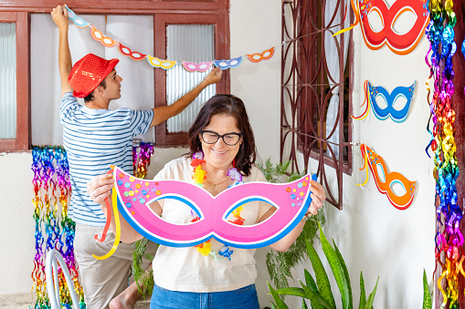 Brazilian mother and adult son joyfully prepare their home for a lively Carnival celebration, capturing the spirit of family and festivity. They are smiling as they decorate the house wall with carnival masks.