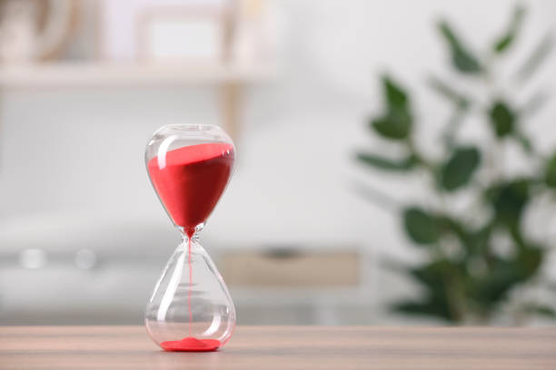 Hourglass with red flowing sand on table against blurred background. Space for text