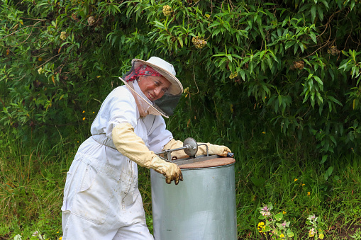 Beekeeper looking at the camera while operating a honey extracting machine. She is dressed in a white protective suit, with a backdrop of forest leaves. The metallic honey extracting machine is visible