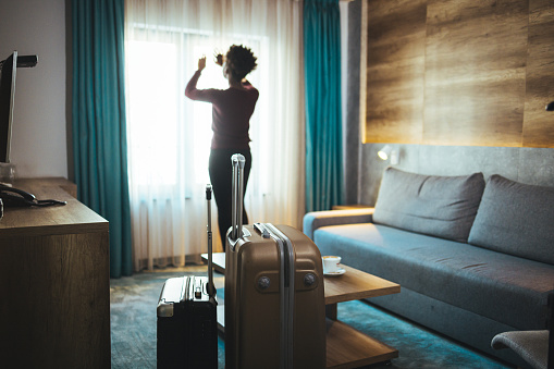 Luggage in modern hotel room with happy young adult female relaxing nearly window, African woman tourist looking to beautiful nature view. Time to travel, relaxation, journey, trip and vacation concepts