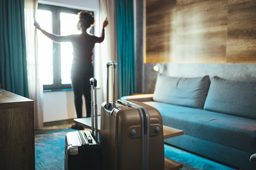 Luggage in modern hotel room with happy young adult female relaxing nearly window, African woman tourist looking to beautiful nature view. Time to travel, relaxation, journey, trip and vacation concepts