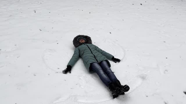 Happy woman lying on snowy lawn at forest and making angel. Young girl playing in snow and enjoying wintertime. Cheerful lady having fun outdoor. Concept of a winter vacation. Close up Slow motion