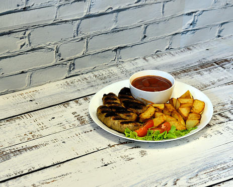Grilled sausages with fried potatoes and tomatoes with lettuce and sauce on a wooden light table. Close-up.