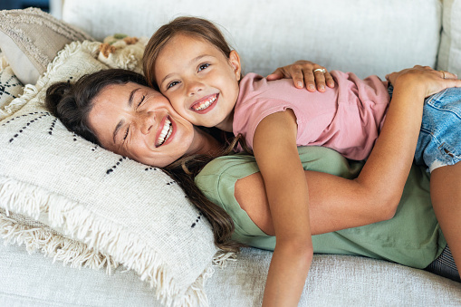 Lovely mother embracing her cute daughter on the sofa at home. Happy mom and little girl laughing, hugging and playful
