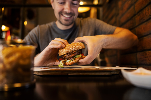 Selective focus on hands holding a hamburger with a hungry man in a blurry background in a fast food restaurant.