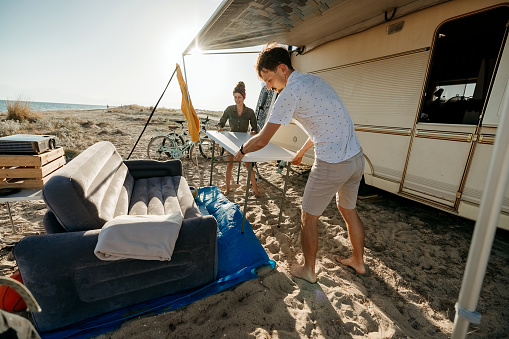 Couple sets up a table and chairs in front of their camper, which is parked not far from the sea