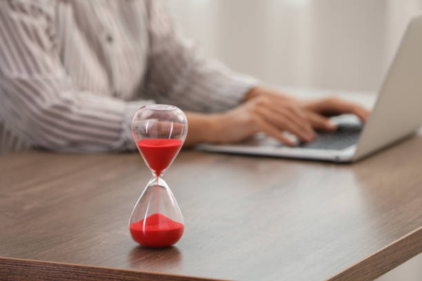 istock Hourglass with red flowing sand on table. Woman using laptop indoors, closeup 1905733532