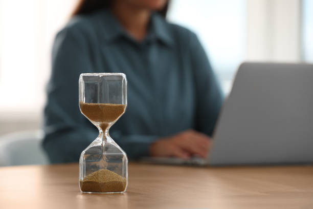 istock Hourglass with flowing sand on table. Woman using laptop indoors, selective focus 1905734096