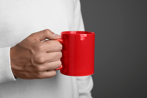 Man holding red mug on grey background, closeup. Mockup for design