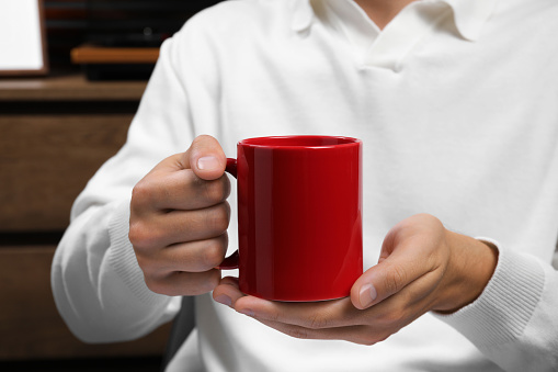 Man holding red mug indoors, closeup. Mockup for design