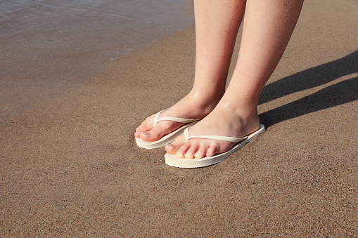 Woman in stylish white flip flops on sandy beach near sea, closeup