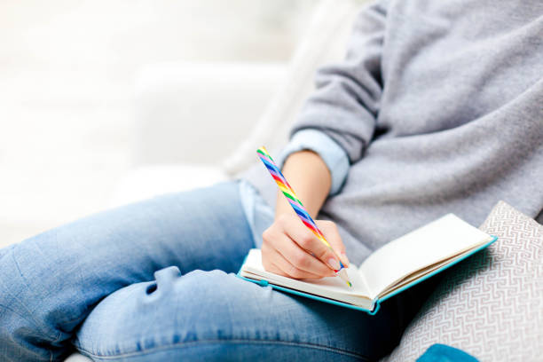 istock Female hand with rainbow pencil is writing goals in notebook. Freelancer is working in cozy place. Girl is planning and dreaming. Young woman is sitting on comfortable sofa. Close up. 1905748659
