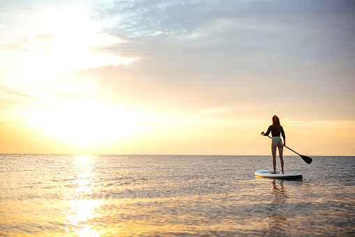 Woman stands firmly on inflatable sup board and floating through sea waves on bright sunset landscape on background