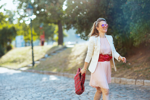 smiling stylish middle aged woman in pink dress and white jacket in the city with red bag and sunglasses walking.