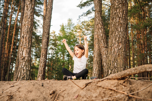 Adorable and happy female kid with open mouth sitting on ground near trees in forest with raised arms. Little sporty girl with dark hair enjoying spending active time in nature.