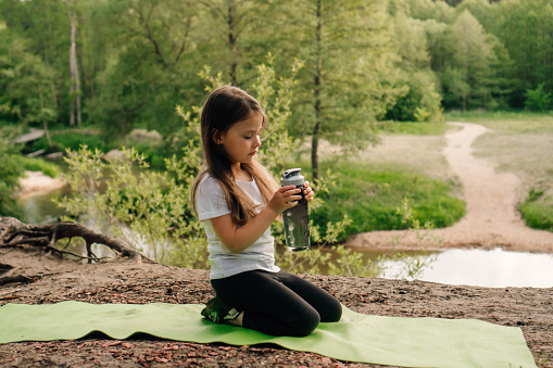 Charming little lady in white t-shirt and black leggings sitting on green sporty mat outdoor and holding bottle of water in hands. Athletic child looking at flask, resting on hill in front of river.