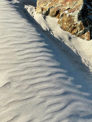 This photograph shows a sandy dune with a large rock, characterized by shadowed ripples created by wind patterns.