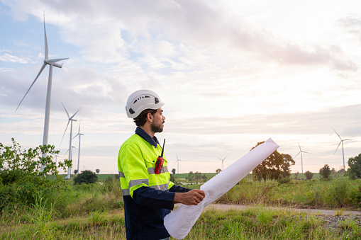 Engineer wearing uniform inspection and survey work in wind turbine farms rotation to generate electricity energy. Maintenance engineer working in wind turbine farm at sunset.