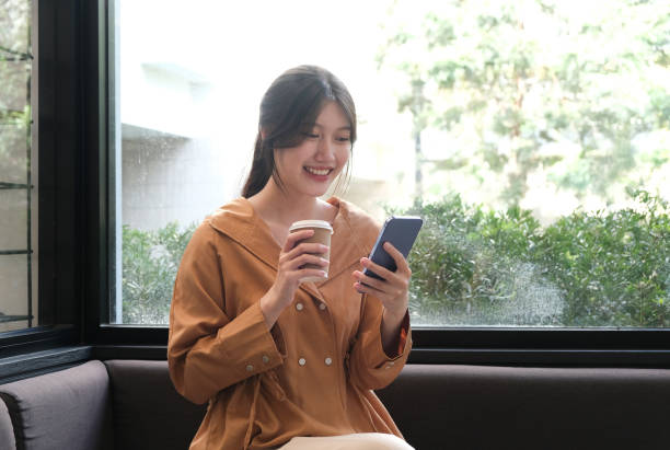 istock Portrait of cheerful young female holding coffee cup and using mobile mobile while sitting in coffee shop 1906131099