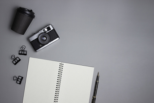 A white notebook with camera, pen, coffee cup and clips over the grey background.