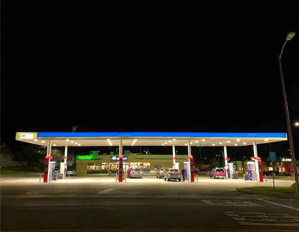This photograph depicts a brightly lit gas station at night, with vehicles refueling and a convenience store visible in the background.