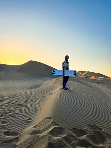 Full body shot of young man standing with sandboard at sunset on top of dune in Huacachina, Ica, Perú