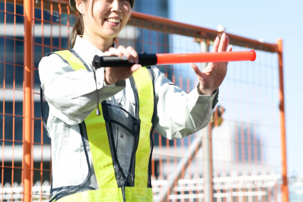a young asian woman of security guard wearing work clothes and a construction helmet and holding a guide light at a construction site - 警備員 日本人 ストックフォトと画像