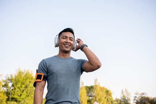 A happy and fresh Asian man in sportswear is enjoying listening to music on his wireless headphone while running in a park in the morning.