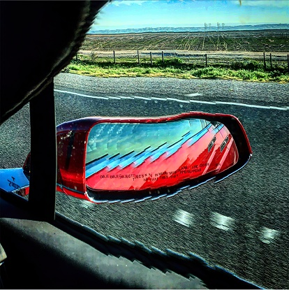 This photograph shows a red car's side view mirror reflecting the road and the surrounding landscape, with a warning text that objects in the mirror are closer than they appear.