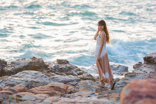 Asian woman standing at the beach in sunset. A woman stands on a rock in the sea during a storm. Tie-dye and long dress, the waves break on the rocks and white spray rises.