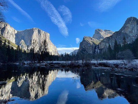 This photograph depicts a tranquil winter scene in Yosemite National Park, showcasing the majestic granite cliffs and their reflections on the still waters of the Merced River.