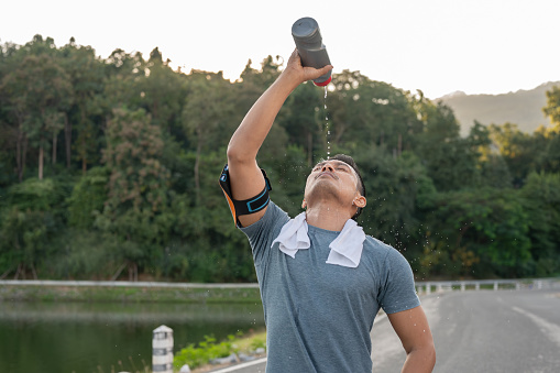 A thirsty, tired Asian man in sportswear is drinking pouring water from a bottle on his face after a long run outdoors in the morning.