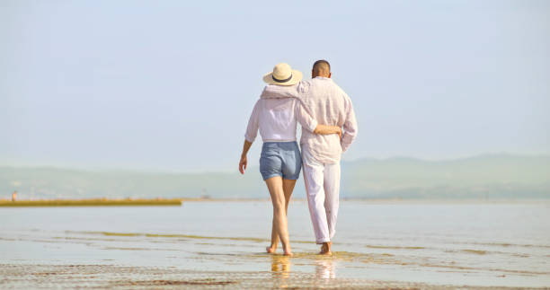 couple walking on beach - women wading sun hat summer foto e immagini stock