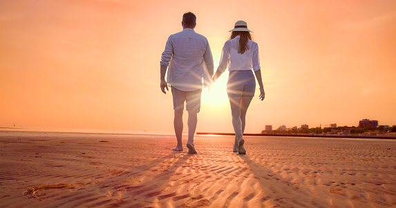 Rear view of couple with holding hands while walking on beach during vacation at sunset.