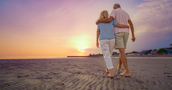 Rear view of senior couple with arm around walking on beach during vacation at sunset.