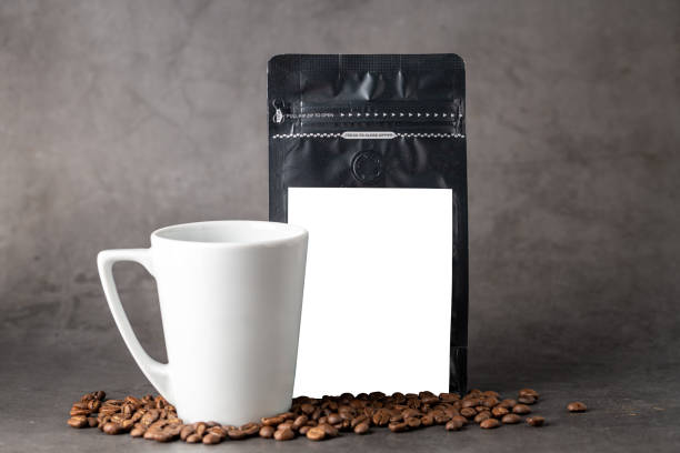 istock Black coffee foil packaging bag and coffee cup with blank white label on stone table 1906352698